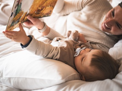 Mamma e bambino si godono le coccole nel letto appena rifatto, con la finestra aperta e i suoni della natura in sottofondo.
