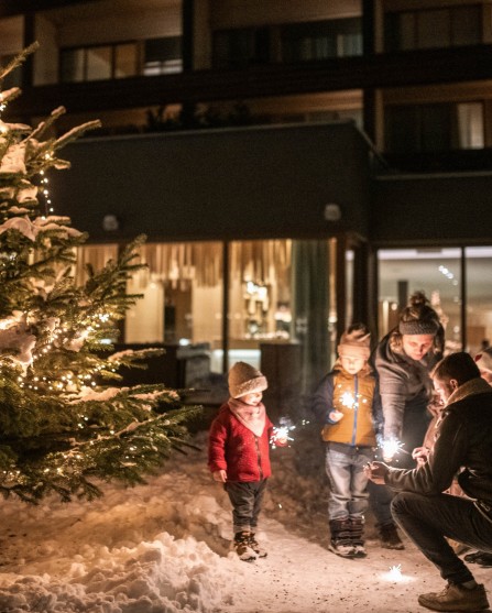 Familie steht vor dem Haus und vor dem Christbaum und zünden einen Sternspritzer an.