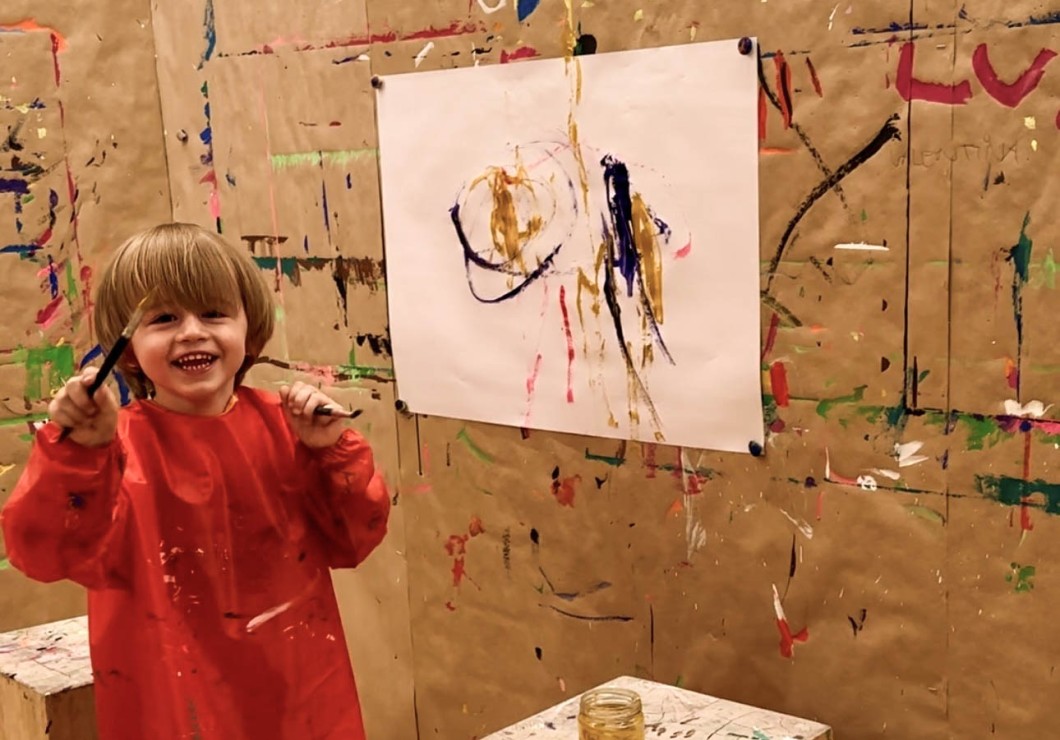 Ein lachendes Kind mit Pinsel in der Hand steht vor einer bemalten Leinwand.