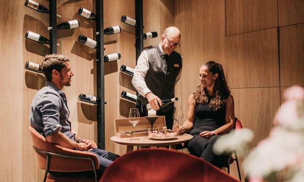 Tre colleghi che discutono intorno a un tavolo nella sala vini del Feuerstein con parete di bottiglie.