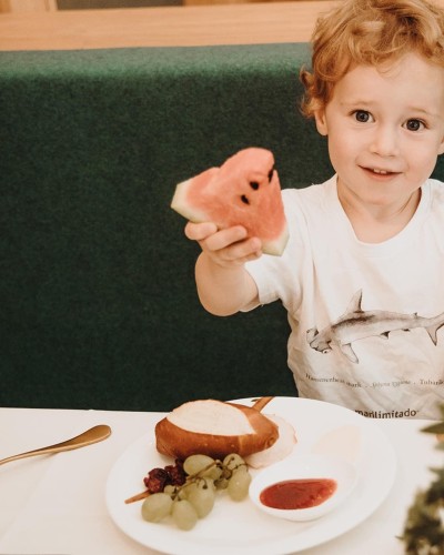 Kleines Kind sitz am Tisch und hält ein Stück Wassermelone in die Luft