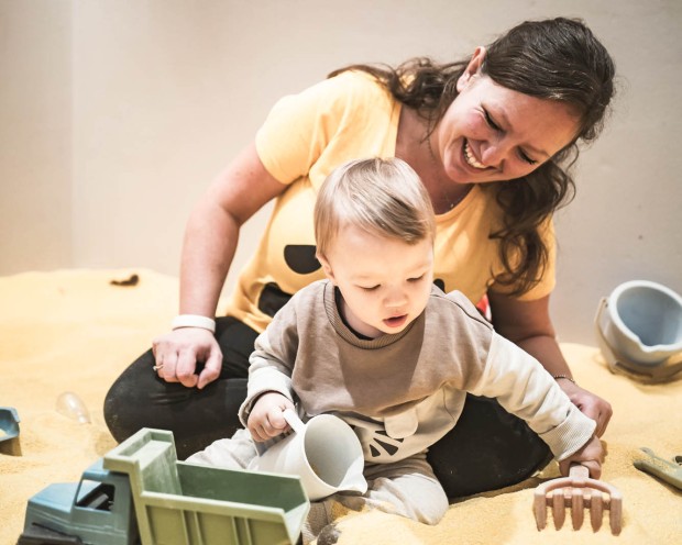 Mamma che gioca con il suo bambino nella sabbiera con camion giocattolo e rastrello.